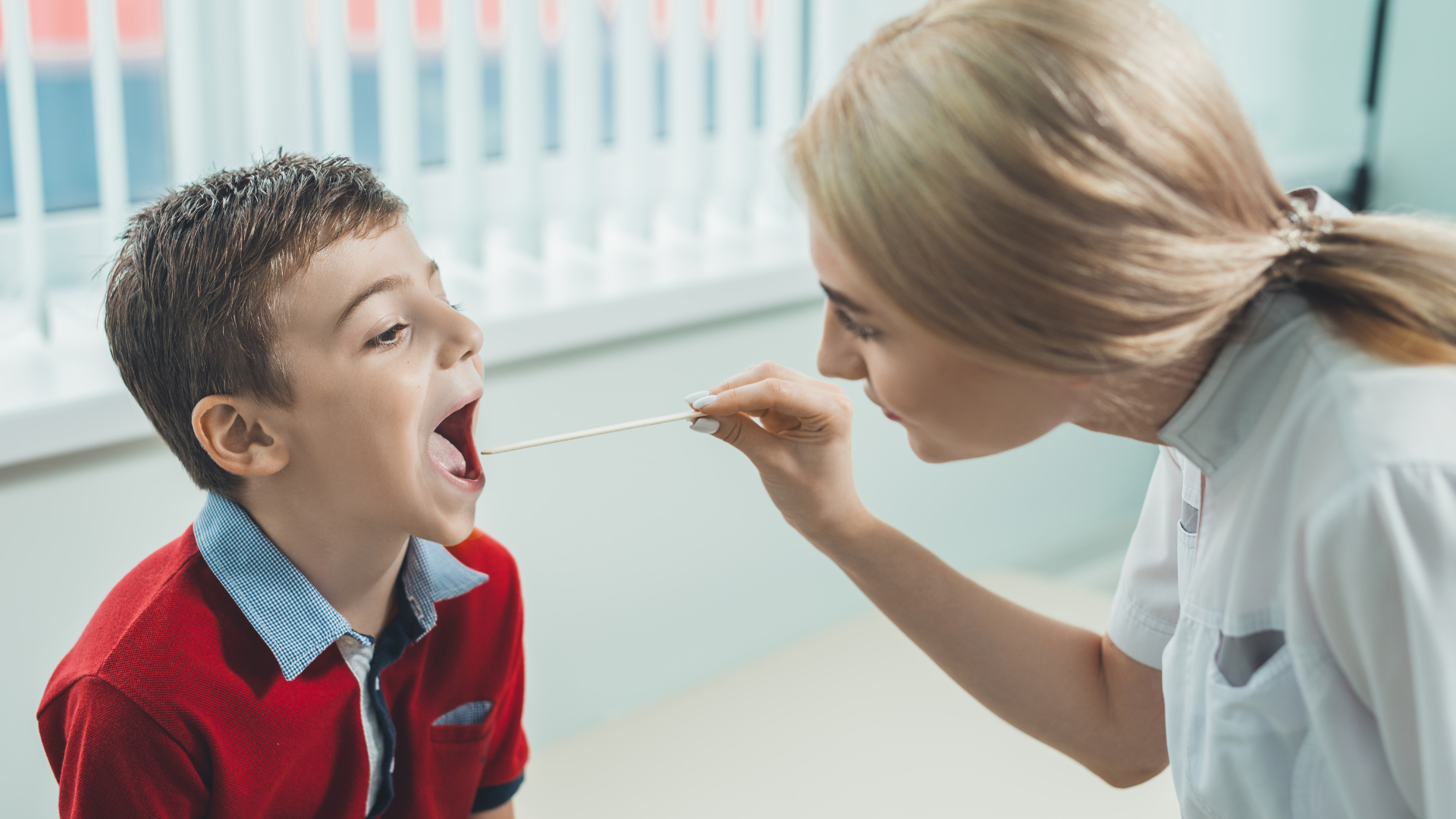 A boy wearing red opening his mouth for a nurse wearing light blue to take a throat swab, testing for strep throat in a traditional manner.