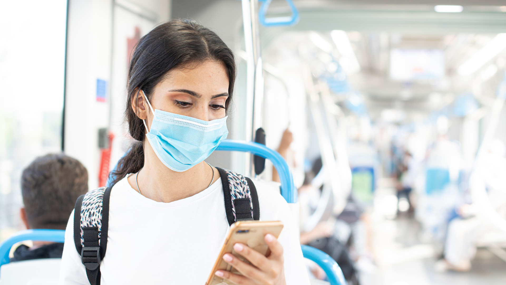 A young woman wearing a medical face mask is sitting in a public transit vehicle. She has dark hair tied back, is wearing a white shirt and a backpack, and is looking down at her smartphone. The background shows other passengers seated, with blue and white interior design elements.
