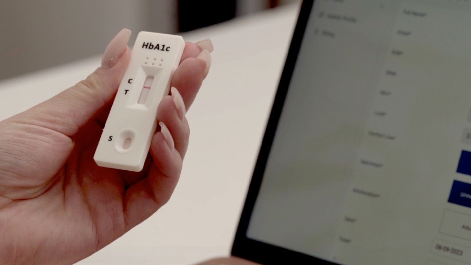 A close-up of a hand holding an HbA1c rapid test device, with a laptop in the background displaying data on the screen.