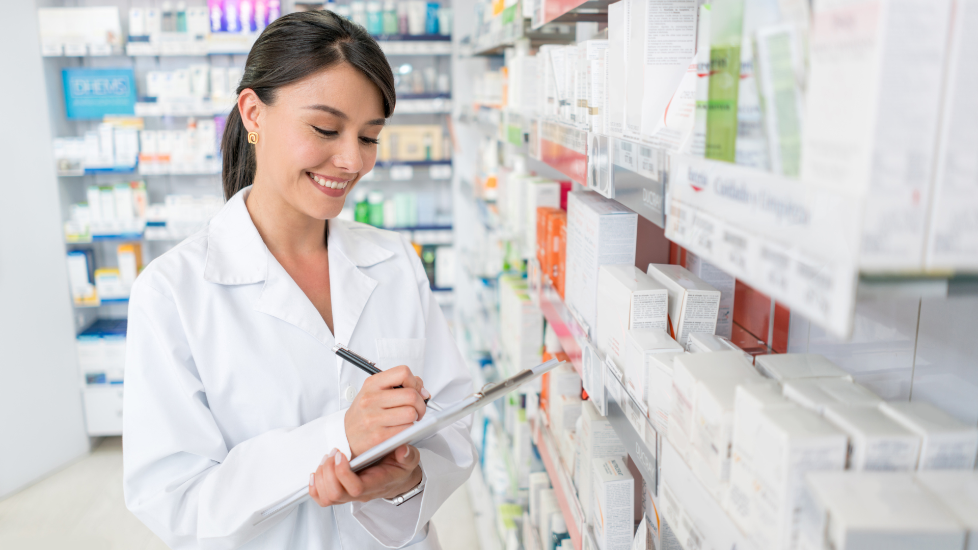 A smiling female pharmacist in a white lab coat is taking notes on a clipboard while standing in an aisle lined with various medical products and medications in a well-lit pharmacy.