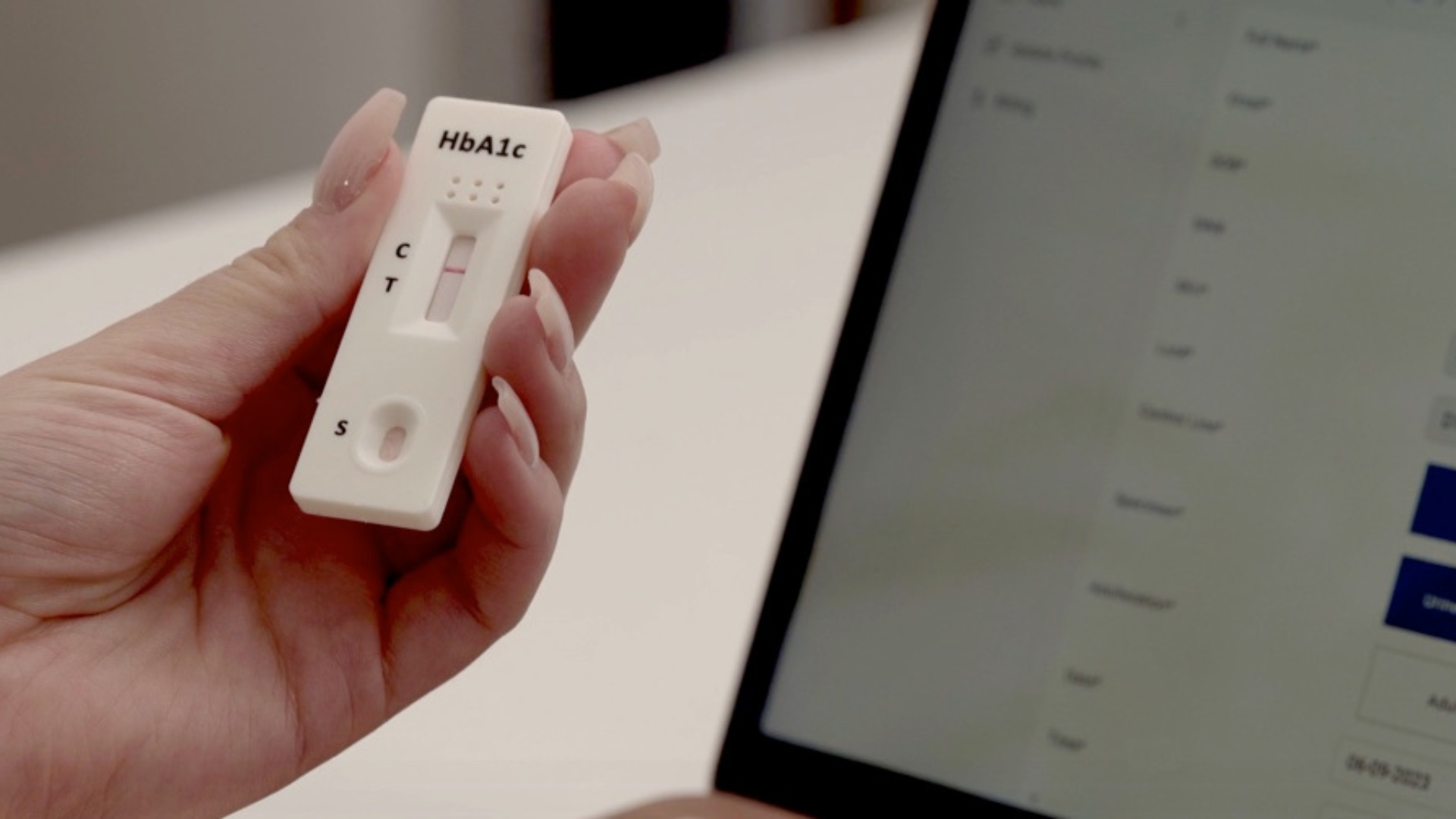 A close-up of a hand holding an HbA1c rapid test device, with a visible result window showing test lines. In the background, a computer screen is partially visible, indicating a clinical or laboratory setting.