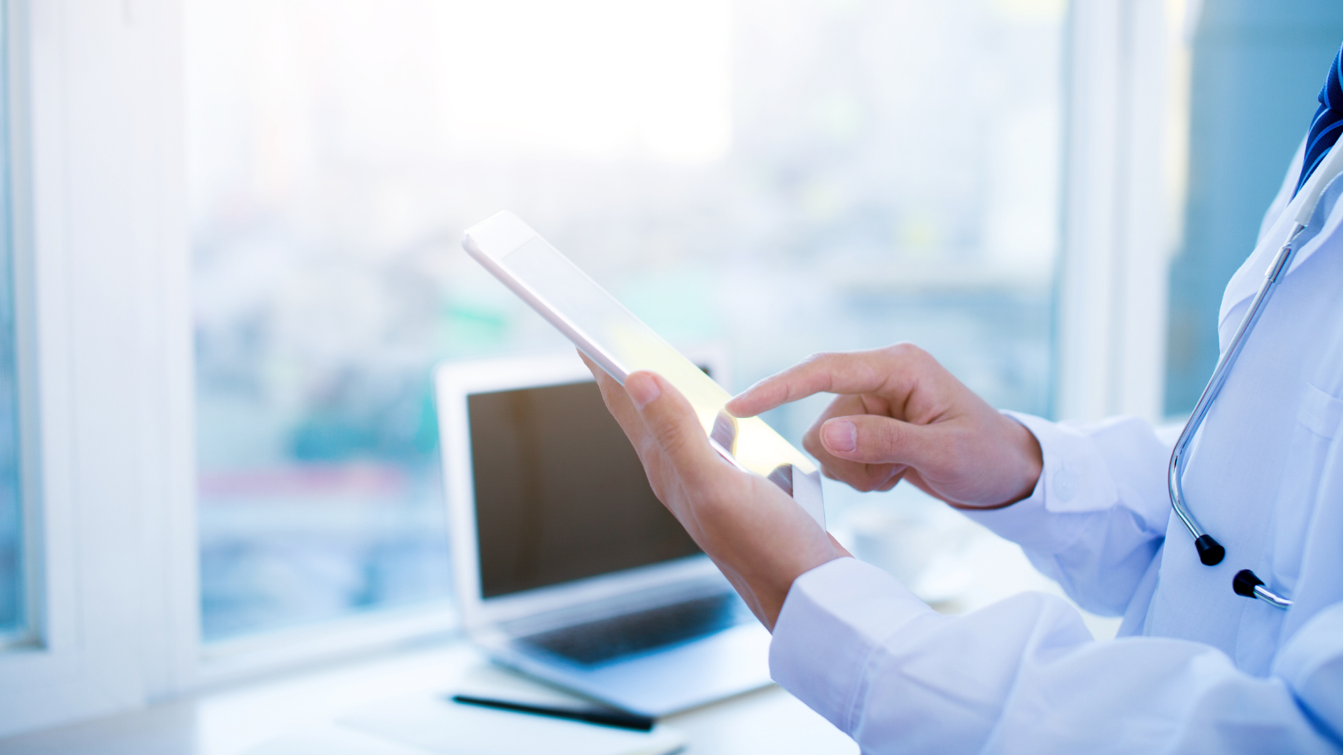 Close-up of a healthcare professional using a tablet with a stethoscope draped over the shoulder, symbolizing the use of advanced medical diagnostics and digital healthcare solutions. A laptop is visible in the background, indicating a focus on integrating technology in healthcare settings.